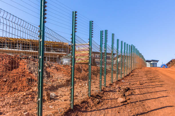 New Security electrical wired fence alongside dirt road with building construction of countryside living apartments in progress with blue sky